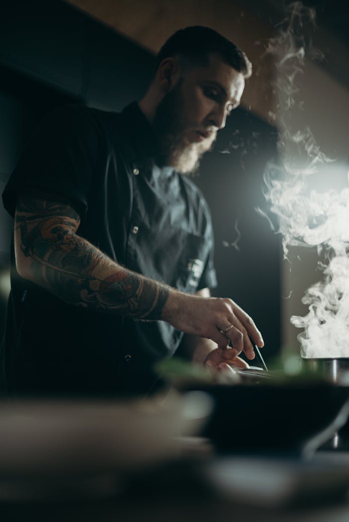 Tattooed chef prepares a dish with focus, surrounded by steam.